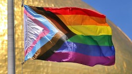 Image of the Pride Progress flag, with the brown, black, pink, and blue chevron, flying over a blue sky.